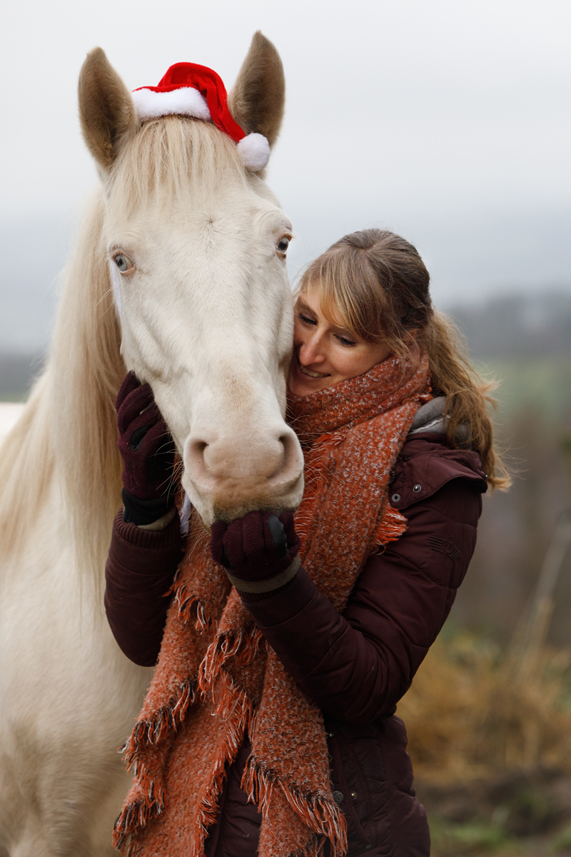 Ein helles Pferd mit einer Niklausmütze und eine junge Frau kuscheln. 